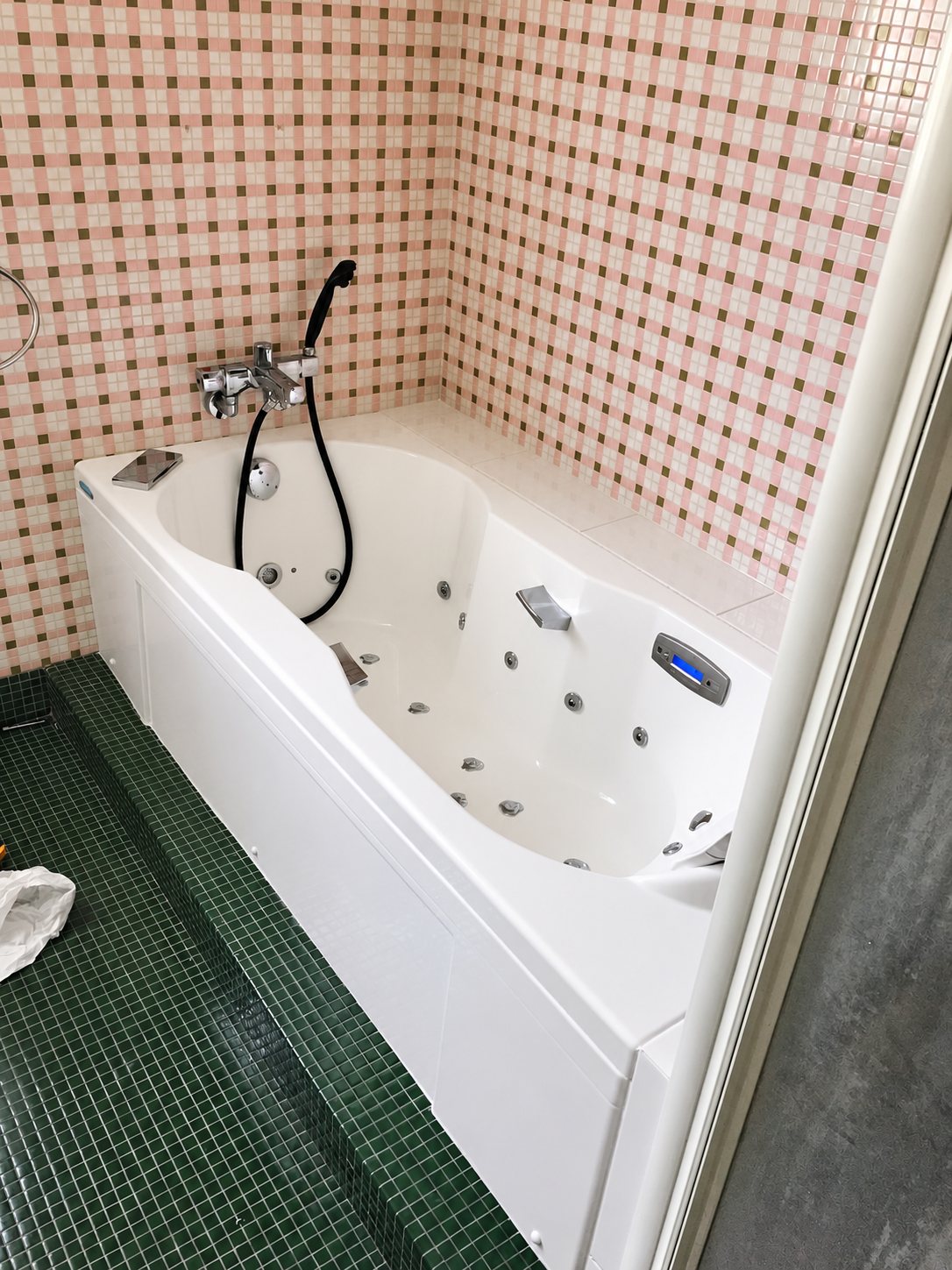 Une baignoire balnéo blanche avec jets et mitigeur chromé, encastrée dans une salle de bain aux murs en mosaïque rose et au sol en mosaïque verte.
