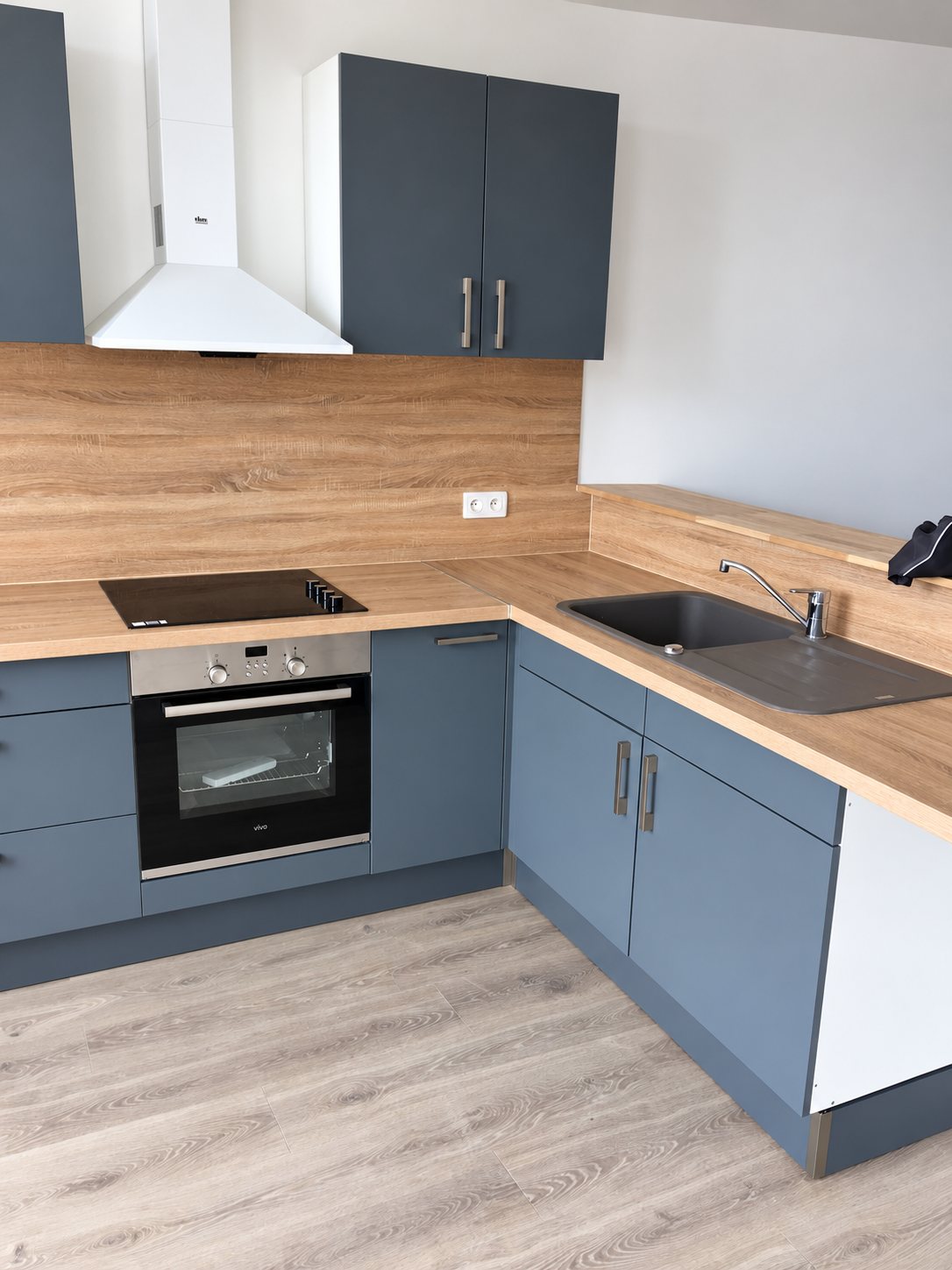 Cuisine neuve en angle avec meubles gris-bleu, plan de travail et crédence en bois, four encastré, plaque de cuisson, évier noir et hotte blanche.